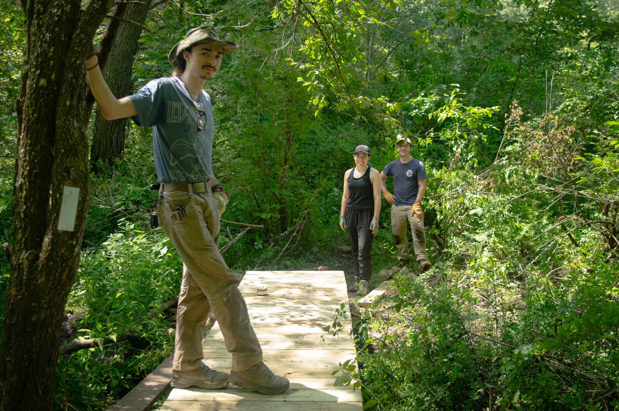 Three people in casual attire, one at the forefront and two others behind, are standing on a narrow wooden bridge surrounded by lush greenery in a forest setting.