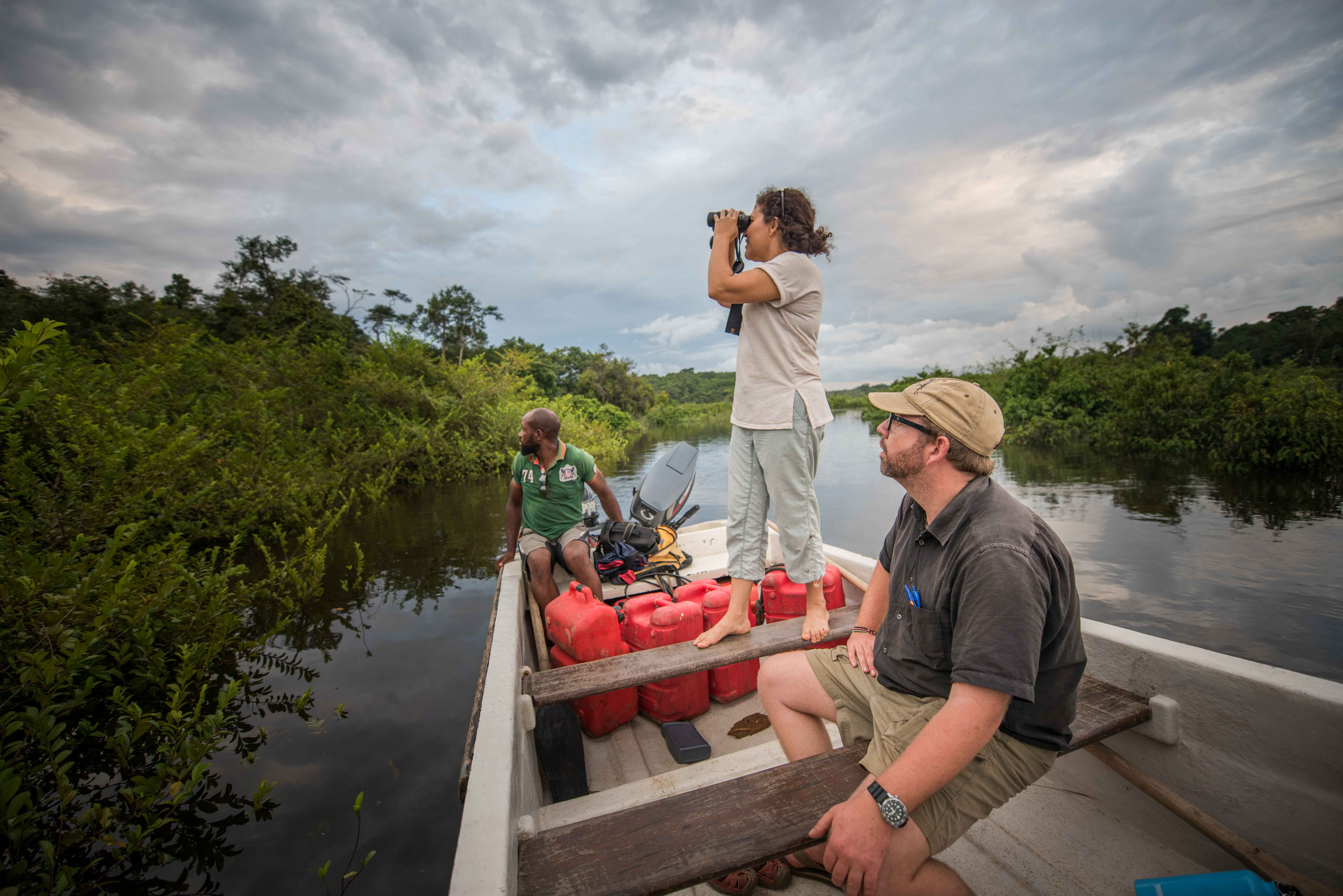 Three people on a boat on a river, one person is standing looking out into the distance with binoculars.