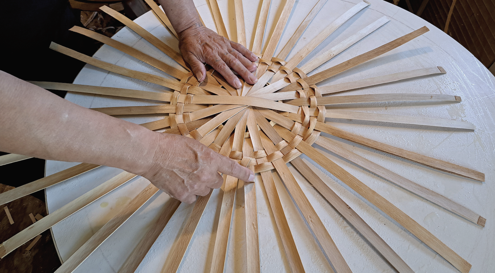 The circular base of a basket with prongs extending. A person's hands are weaving these prongs into the circle. 