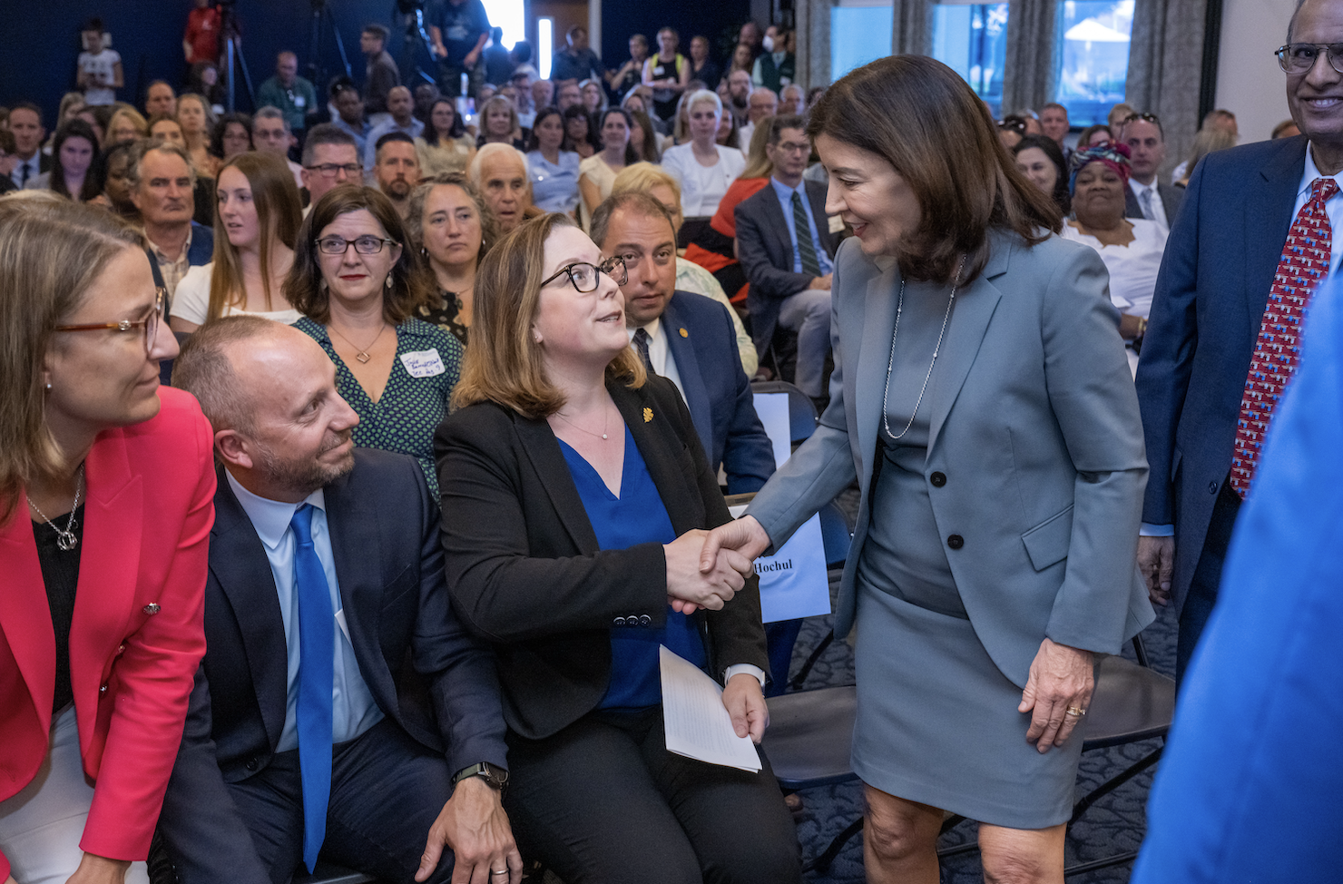 Two women in suits shake hands in the foreground, while a group of other professionally-dressed people sit in the background.