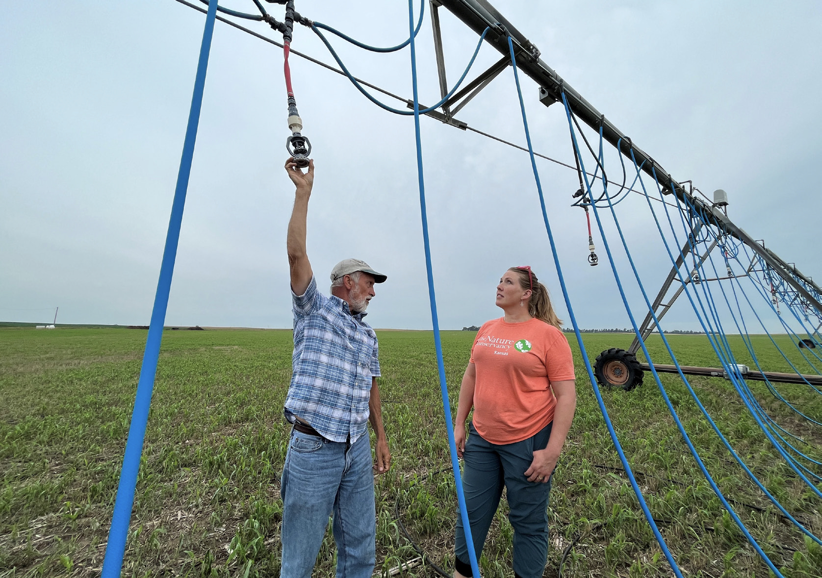 A woman and a man stand in the middle of a field, engaged in conversation beneath an irrigation system.