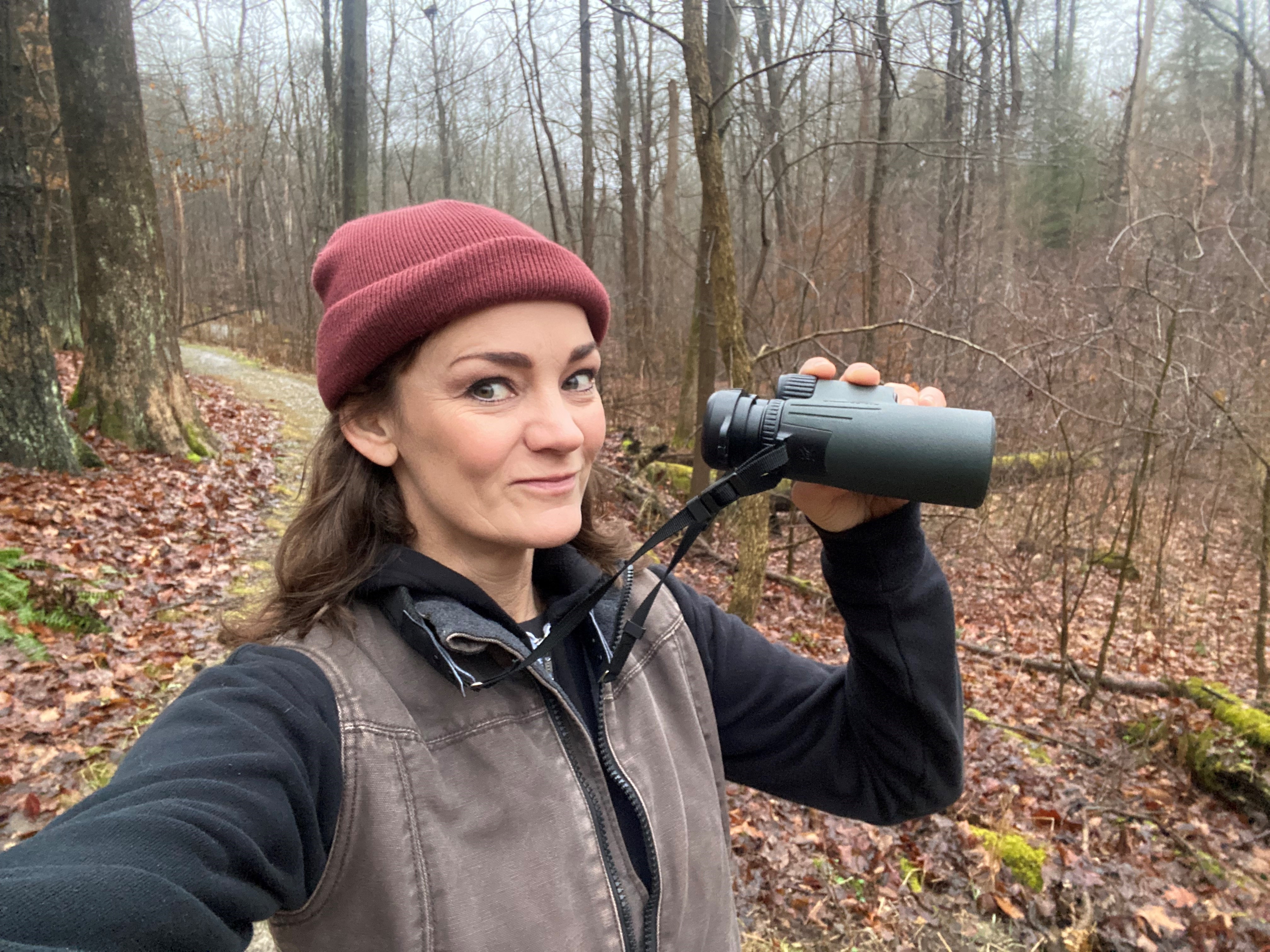 Ann Gilmore holds binoculars in forest.