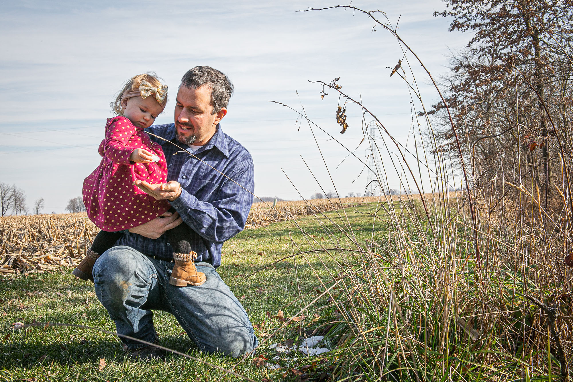 A farmer kneels while holding young girl on knee in a farm field. 