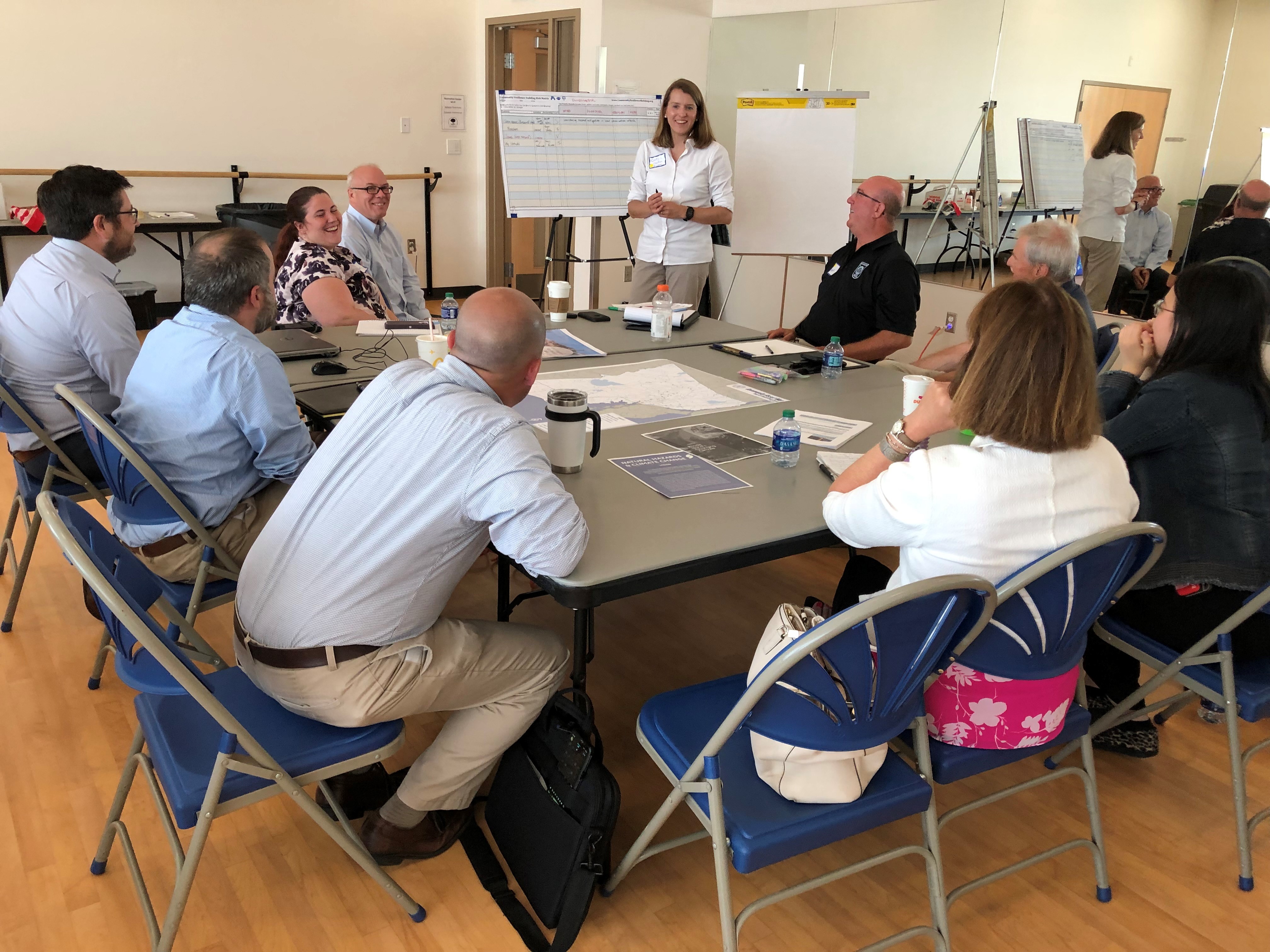 A group of people sit around a table engaged in a meeting. Papers and a map cover the table. A person stands at the front, smiling, beside a large notepad. The setting is a bright room.