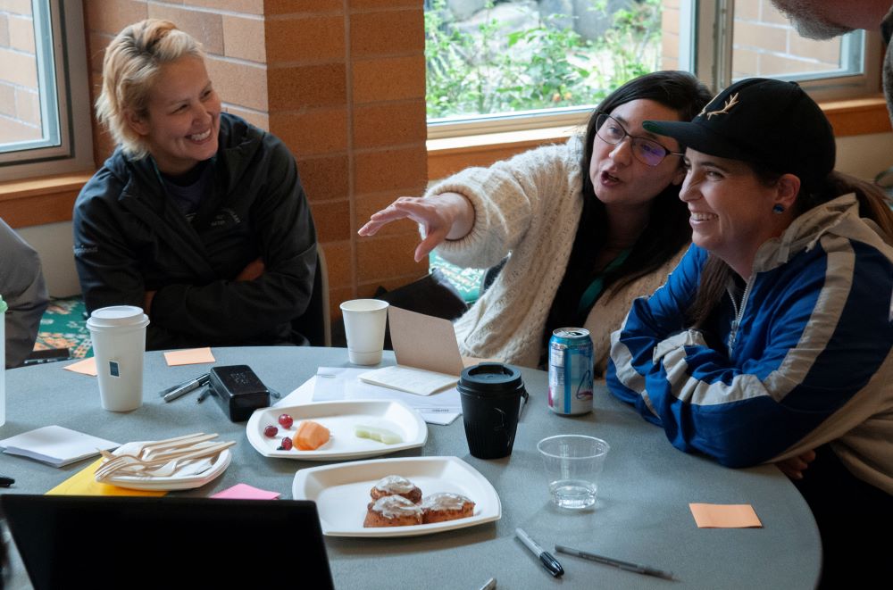 Three people sit together at a table indoors and smile; one person points to something off camera.