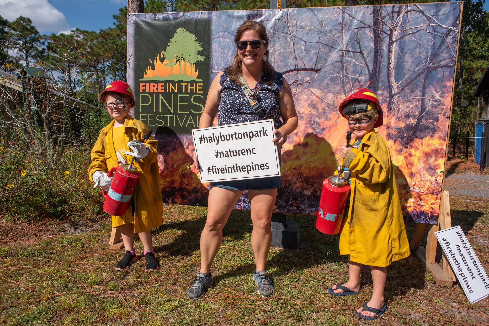 Women and kids posing  in front of fire in the pines sign. Kids are dressed up as firefighters and  holding drip torches.
