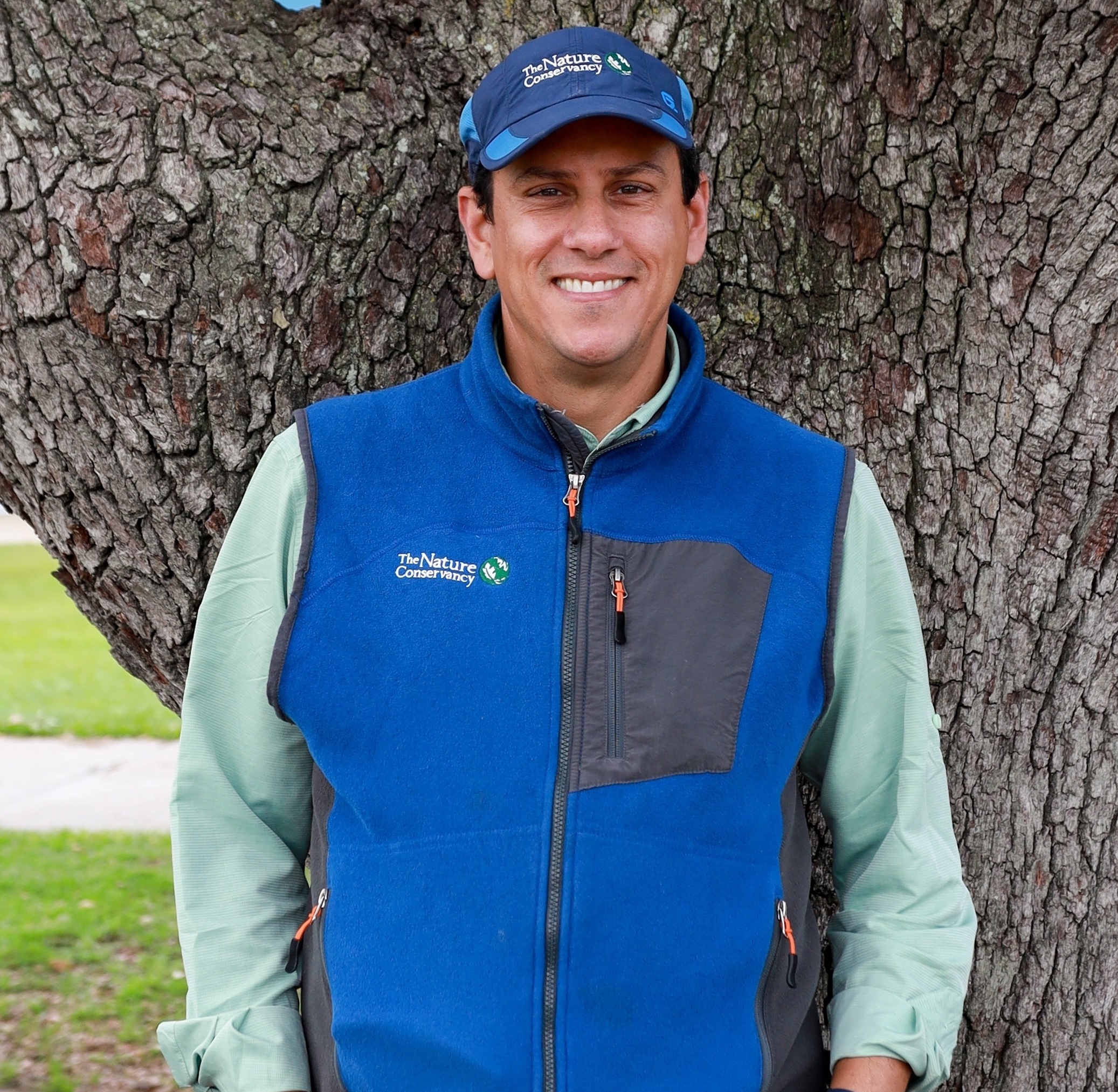A man in a hat stands in front of a tree trunk.