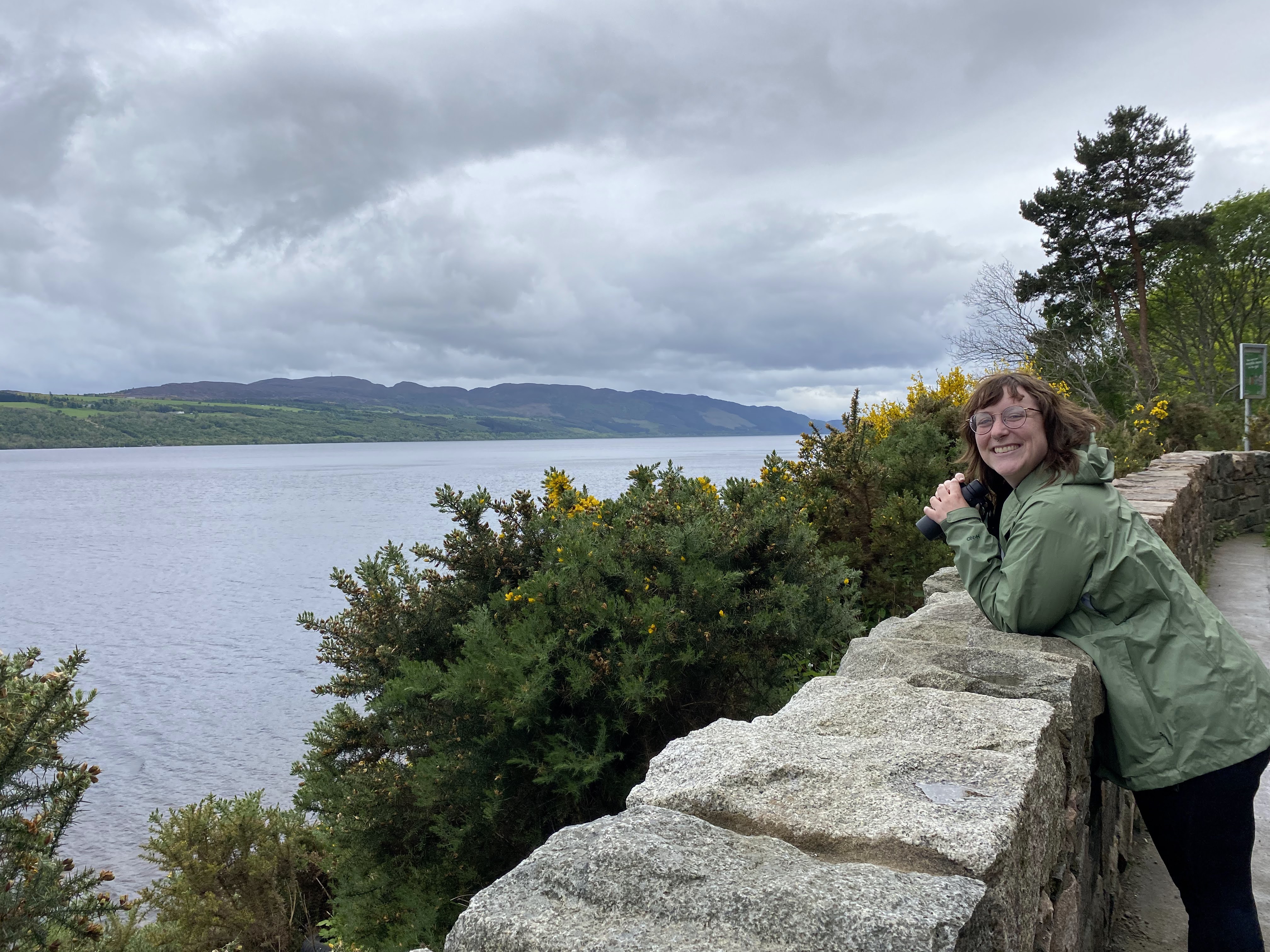A person holding binoculars stands in the forest and looks at the camera.