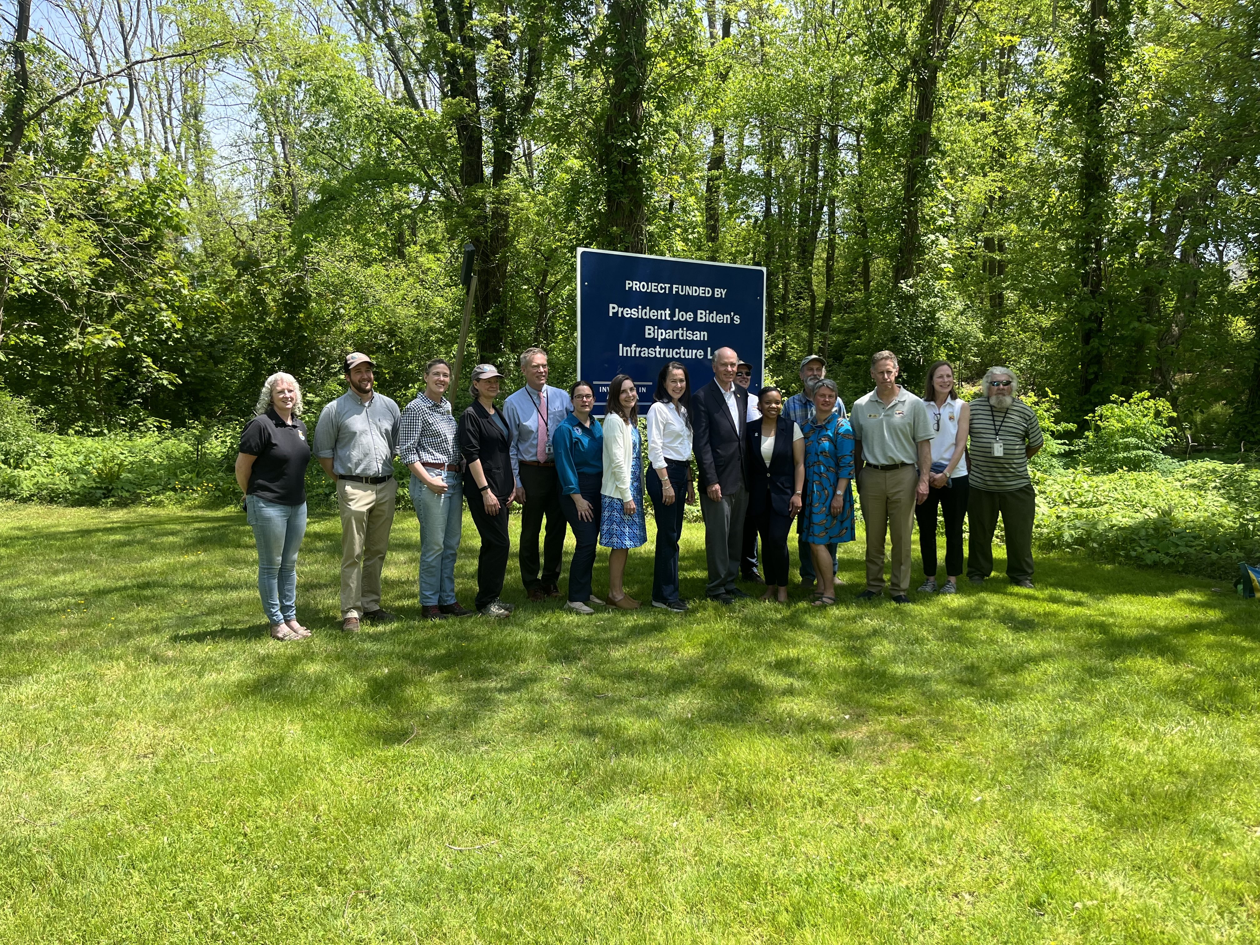 A group of project partners poses for a photo in front of a sign at the High St Dam removal project indicating it was funded by the Bipartisan Infrastructure Law.