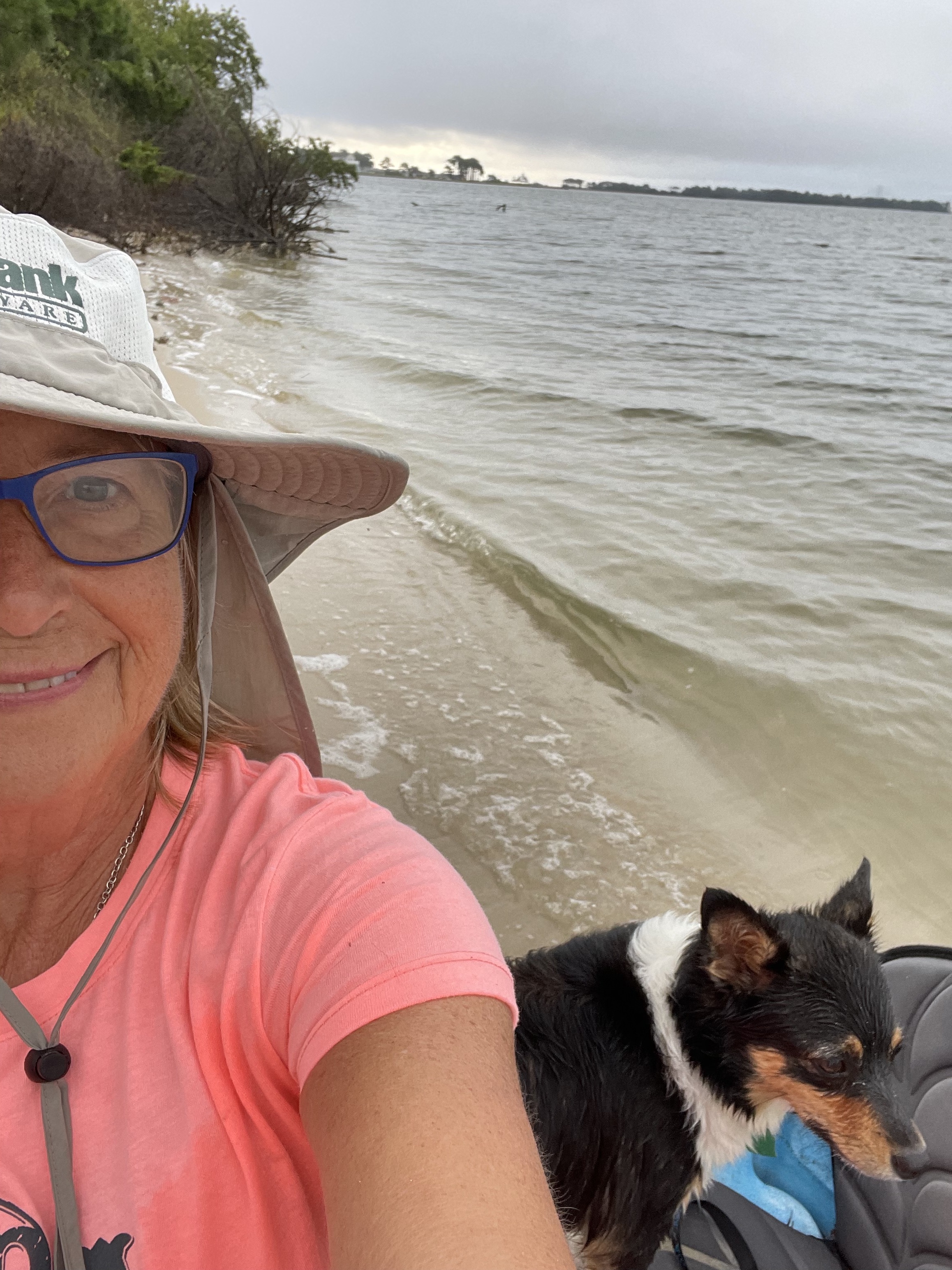 A selfie-style photo of a person smiling at the camera with a dog in front of a beach.