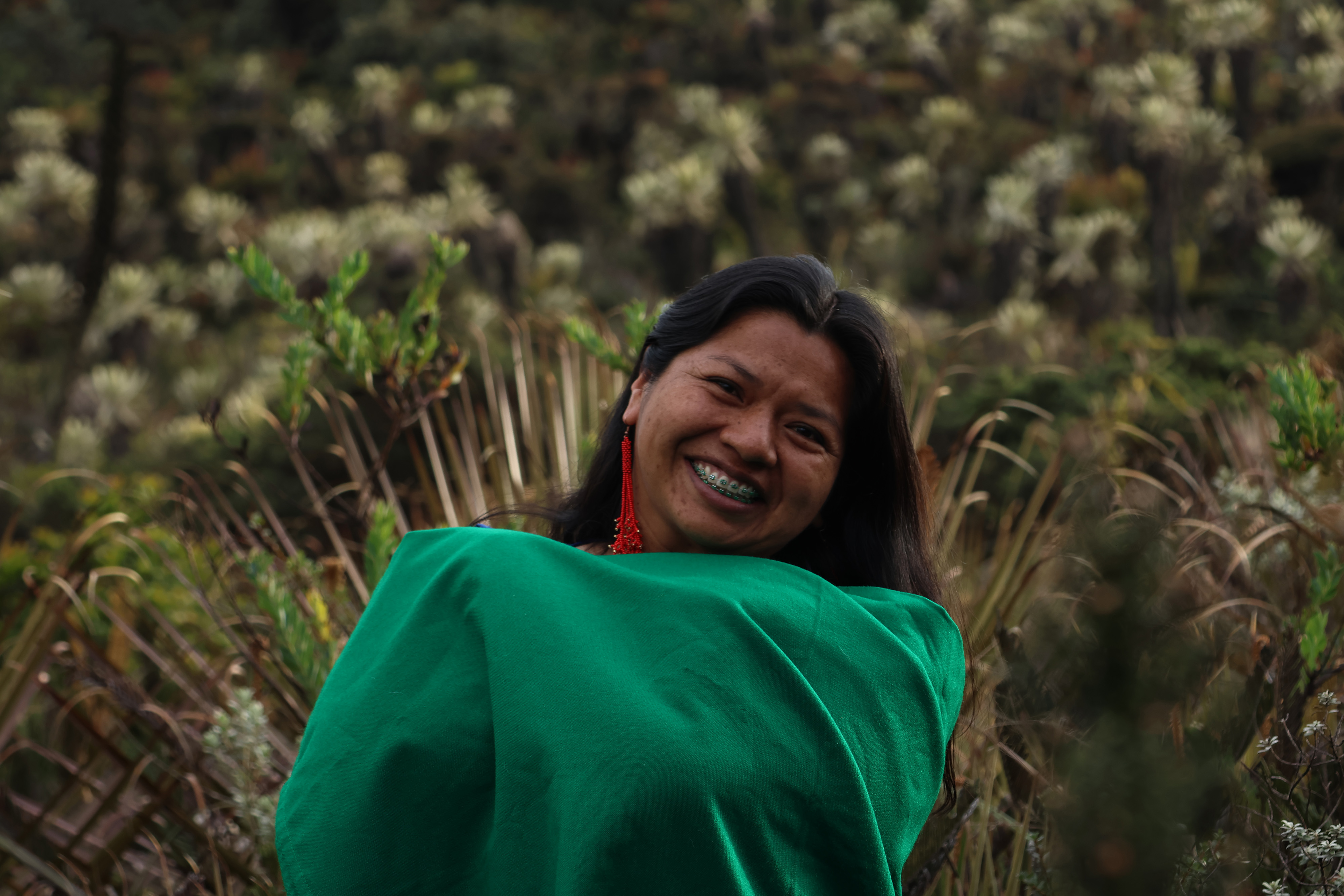 Portrait of Eliana Muchachasoy, wearing an emerald green scarf and red earrings, with her dark hair falling over her shoulders and smiling to the camera, with a nature background. 