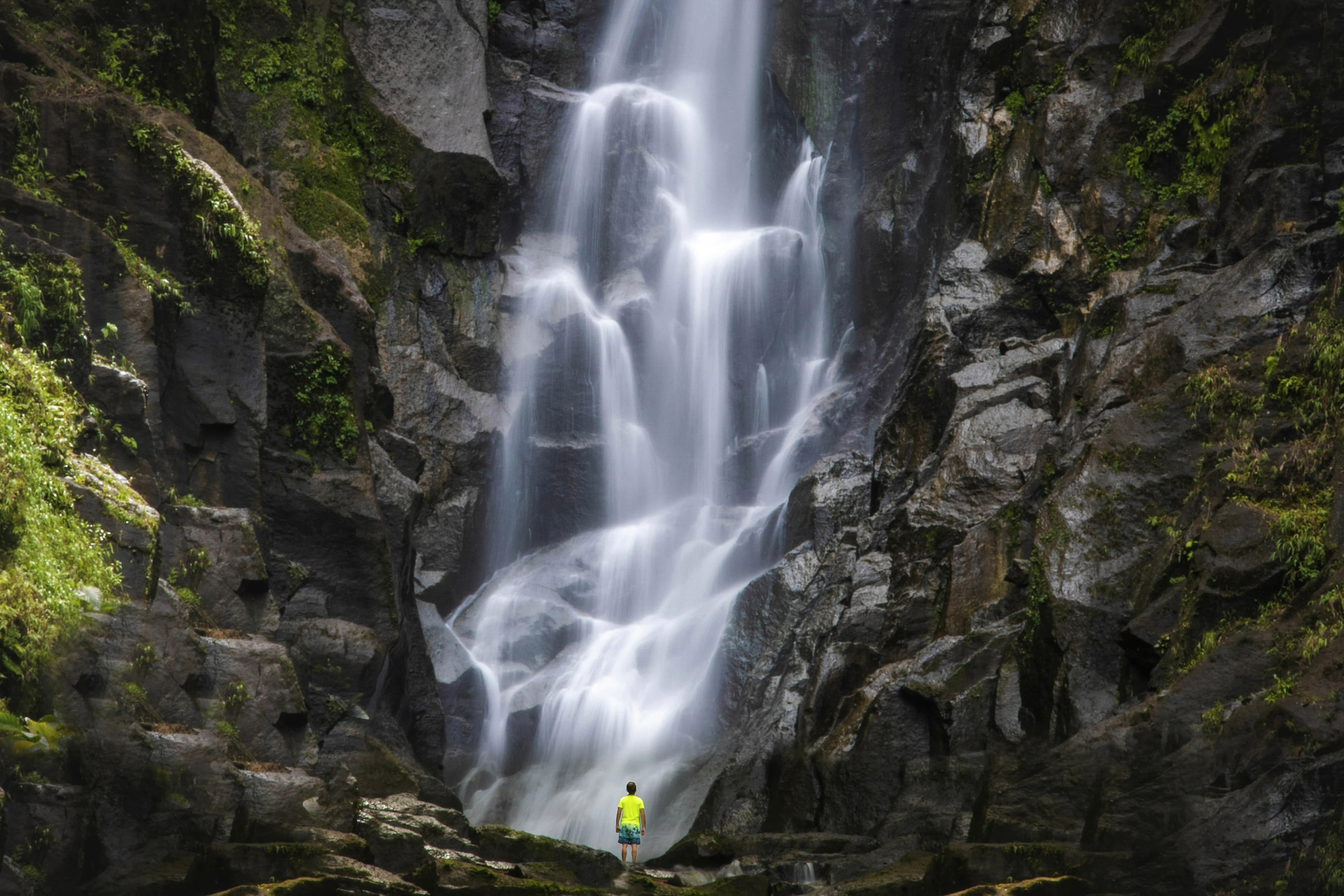 a person in a neon yellow shirt standing at the bottom of a large waterfall, looking up.