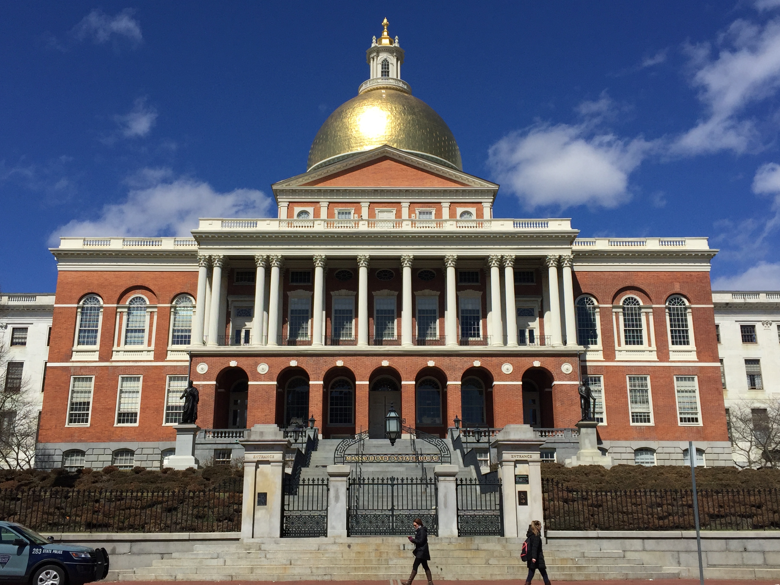 Horiztonal image of the Massachusetts State House with two people walking on the sidewalk in front. The building is mostly brick and has a gold dome on top. 