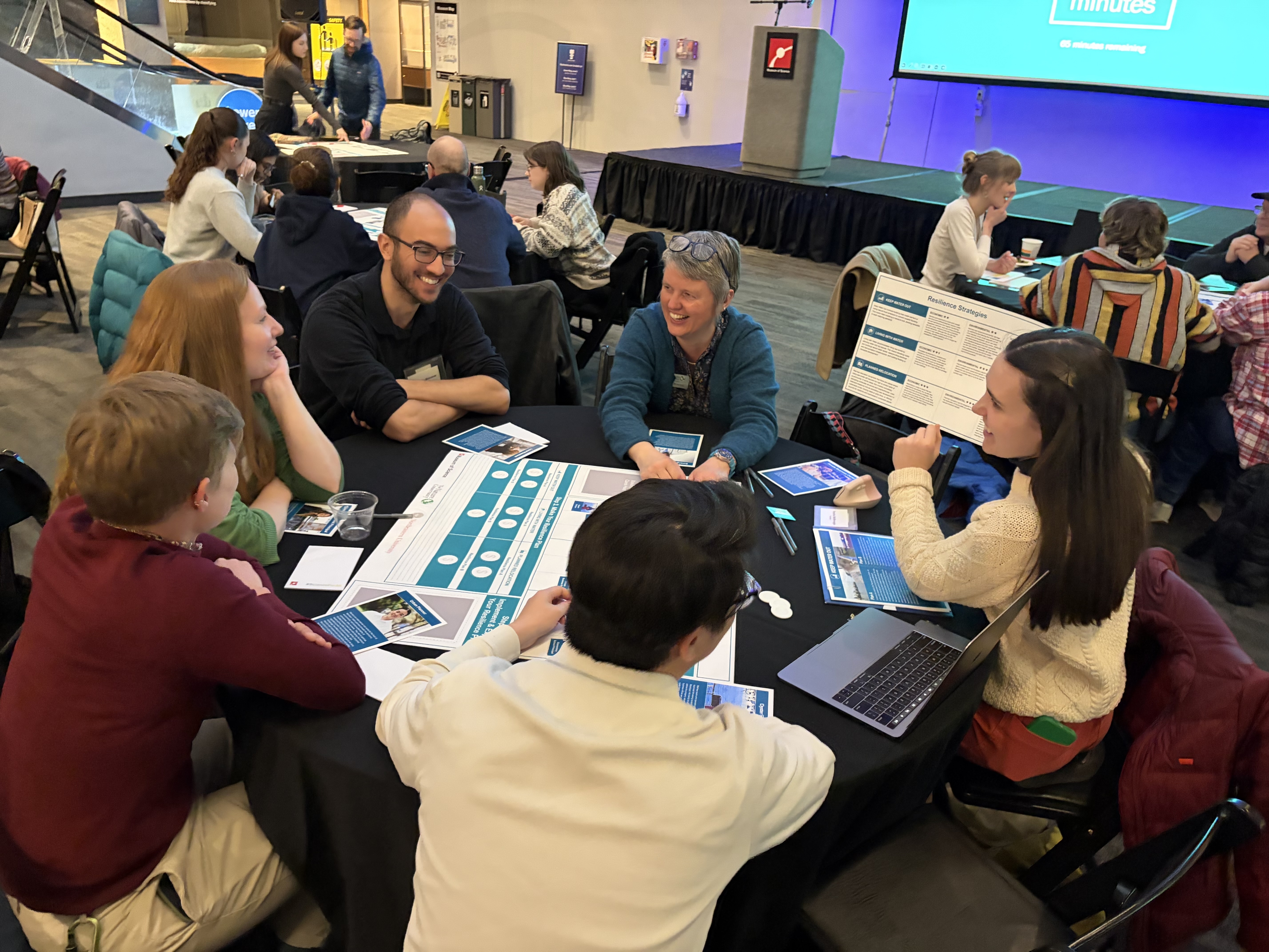A group of people sit around a round table in an auditorium at the Museum of Science. 