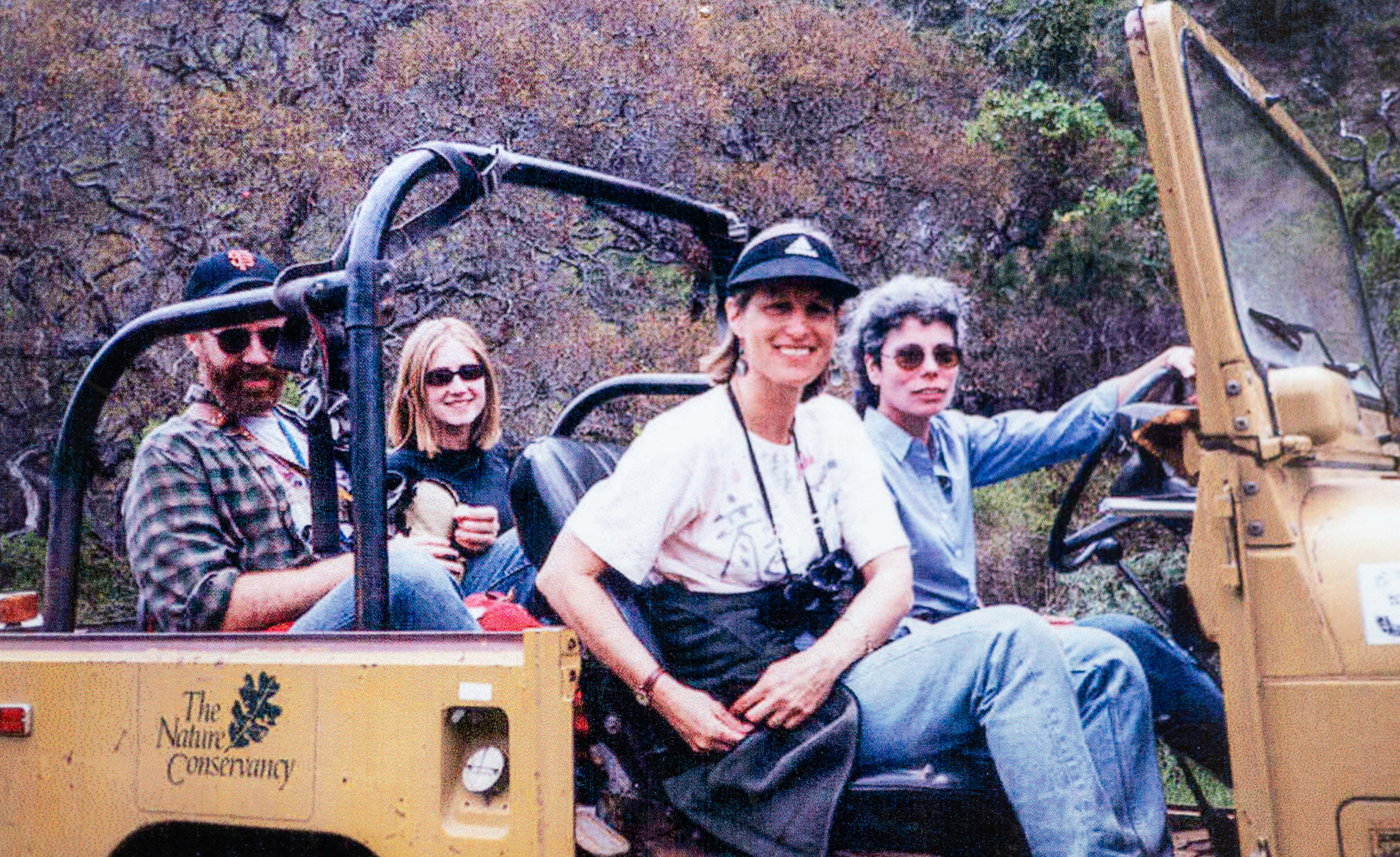 A group of people tour around a natural area in a yellow, open air vehicle. The driver is a woman wearing sunglasses.