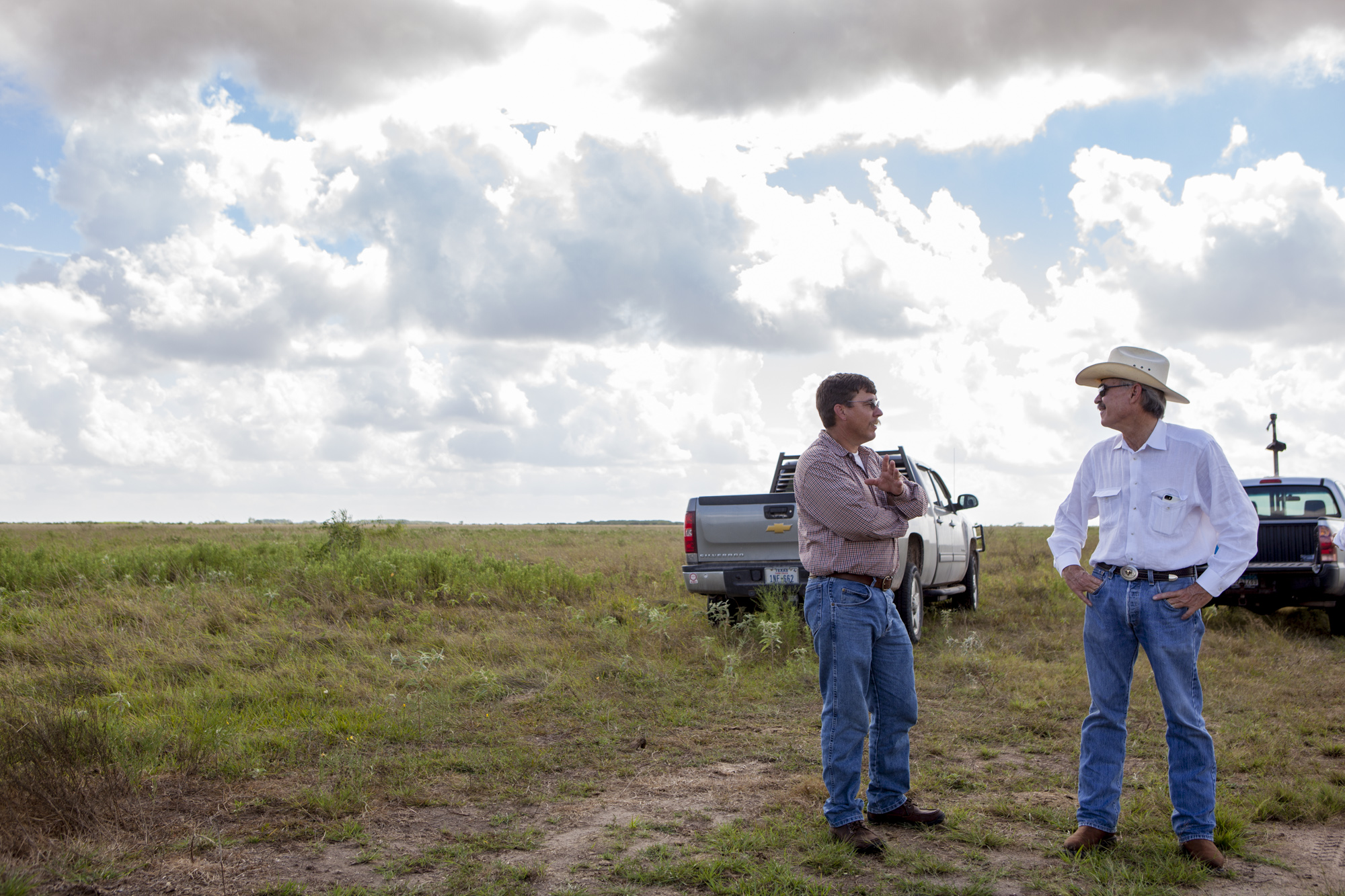 Two men stand among green grass talking animatedly.