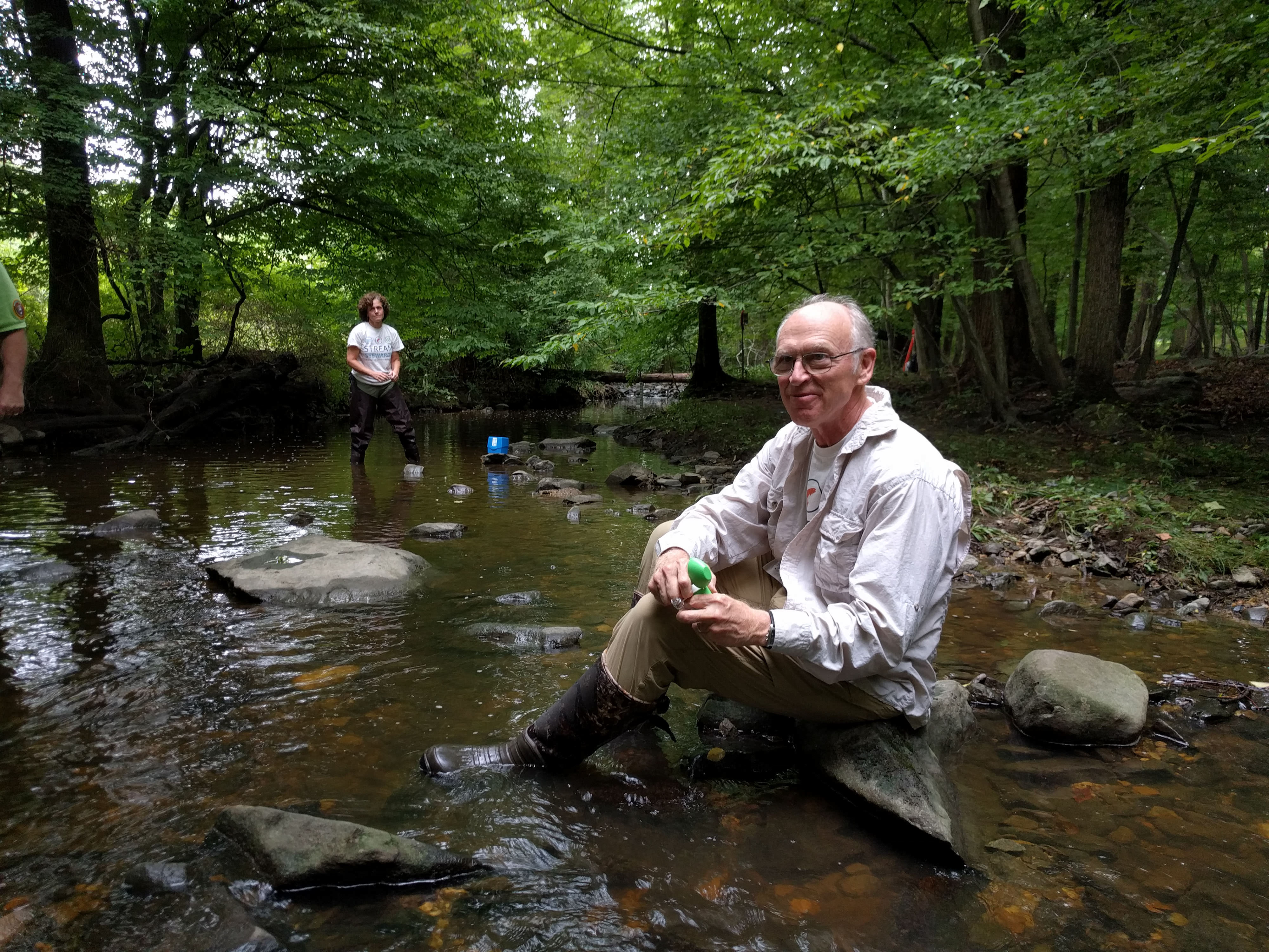 A peron sits on a rock in a shallow river looking into the camera.