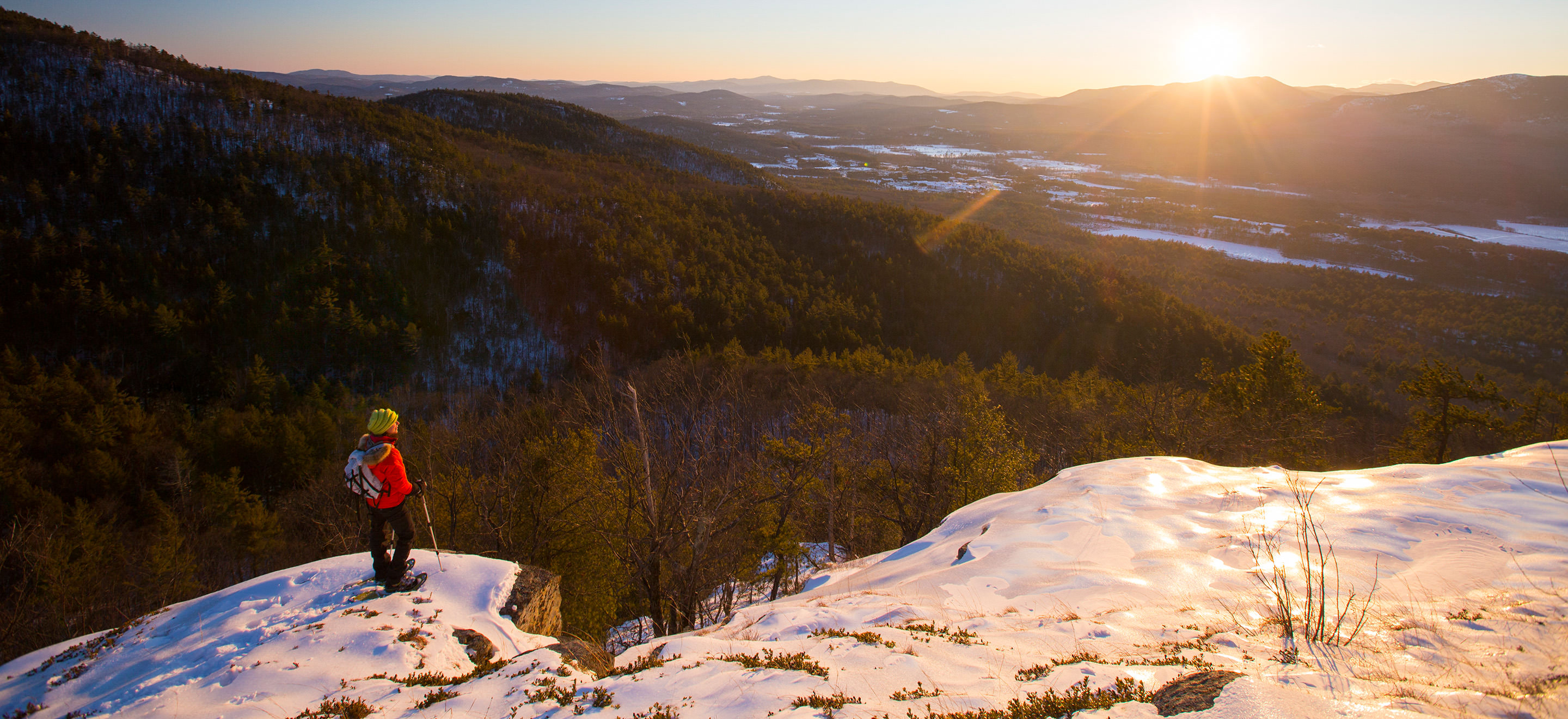 A hiker stops to take in the view of the sun setting over a mountain valley.