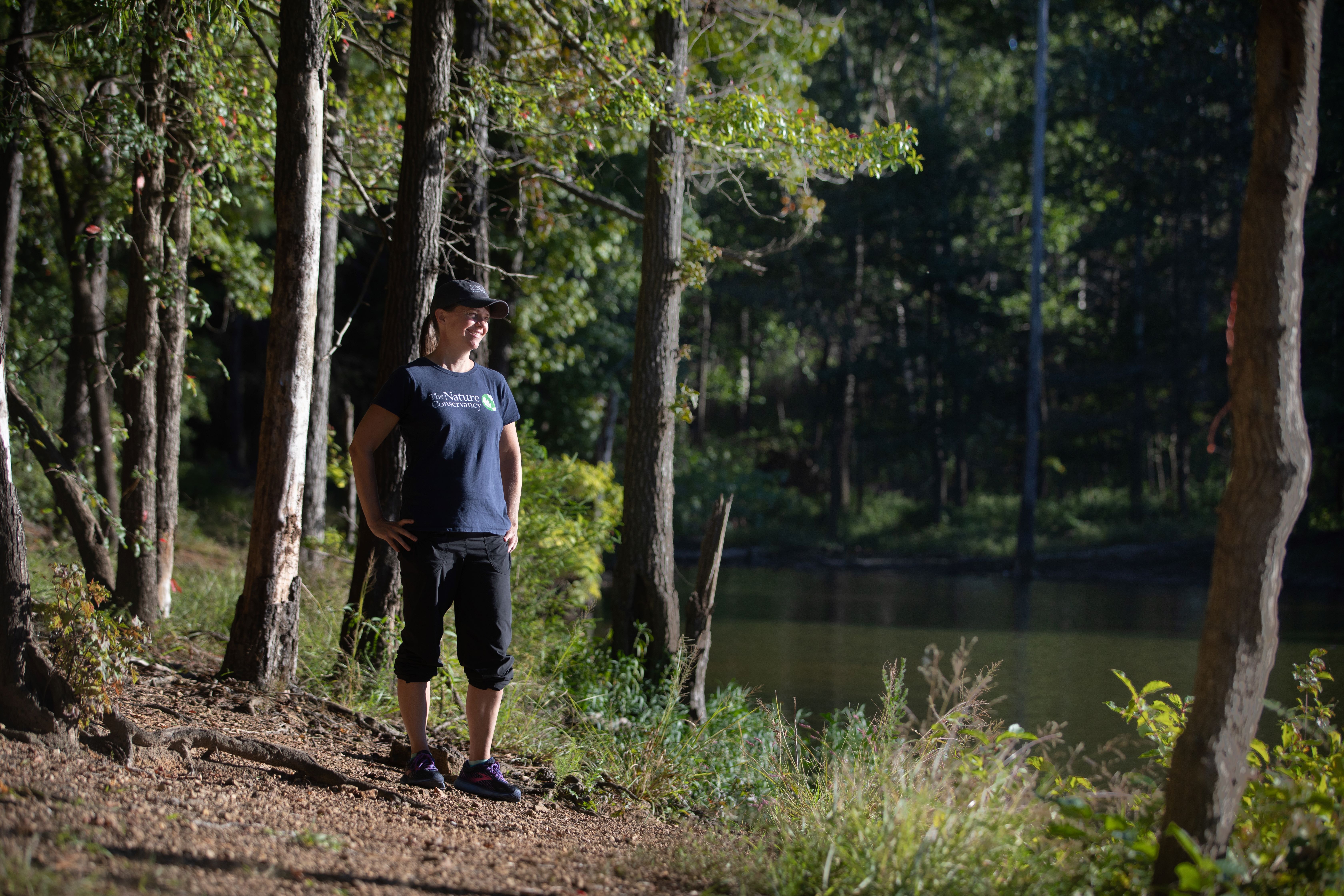 A woman stands at the edge of the forest next to a river.